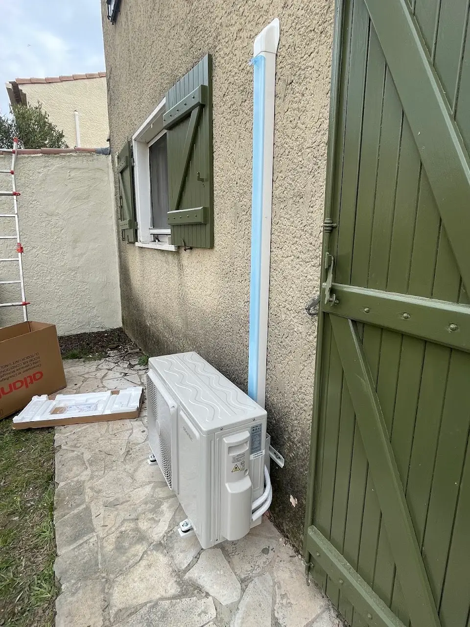 Unité extérieure de climatisation au pied d'une maison aux volets verts